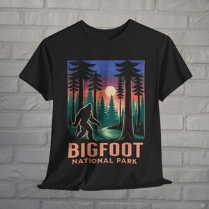 May include: Black t-shirt featuring a Bigfoot graphic in a forest scene with a sunset backdrop. The design includes the text "BIGFOOT NATIONAL PARK" in orange and white. The shirt has a crew neck and short sleeves.
