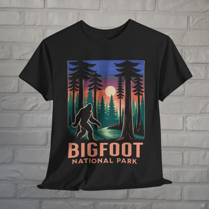 May include: Black t-shirt featuring a Bigfoot graphic in a forest scene with a sunset backdrop. The design includes the text "BIGFOOT NATIONAL PARK" in orange and white. The shirt has a crew neck and short sleeves.