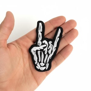 May include: An embroidered patch featuring a white skeleton hand making a rock and roll sign with the index and pinky fingers extended. The design is outlined in black thread, creating a high-contrast graphic element for fabric application.