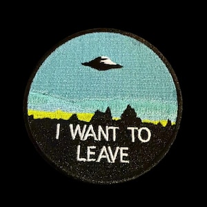 May include: Round embroidered patch with a blue sky, yellow horizon, and black silhouette of mountains. A black and white UFO hovers above the landscape. The text "I WANT TO LEAVE" is embroidered in white.