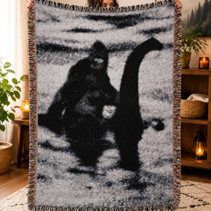 May include: A rectangular woven throw blanket featuring a black and white graphic print of Bigfoot and the Loch Ness Monster standing together. The blanket has a textured, fringed edge with multicolored threads woven into the border trim.