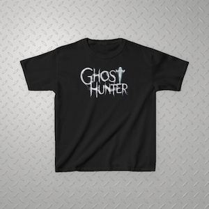 May include: A black short-sleeved t-shirt featuring the white text Ghost Hunter printed on the chest. The lettering has a wispy, ethereal appearance, with the letter T in Ghost replaced by a small, stylized figure resembling a ghost silhouette.