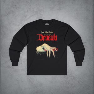 May include: A black long-sleeve t-shirt featuring the text Bram Stoker's Original Dracula in red and white lettering. Below the text is a graphic illustration of a pale, claw-like hand resting on a dark ledge with a red accent underneath.