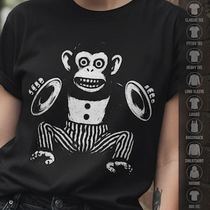 以下が含まれることがあります： シンバルを持った猿の白いグラフィックプリントが施された黒いTシャツ。猿は目を大きく見開いた表情で、2つのボタンが付いたベストとストライプのパンツを着用している。デザインは生地の上でかすれたような質感のある仕上がりになっている。