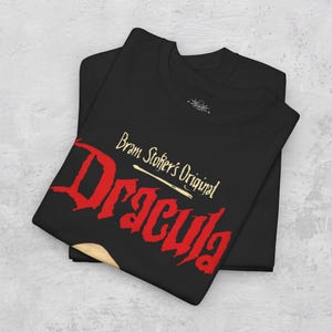 May include: A black t-shirt featuring the text Bram Stoker's Original Dracula in a bold red font with a cream-colored underline. The shirt has a crew neckline and a small graphic logo printed on the inside collar area, showcasing a dark aesthetic.