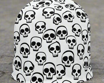 May include: A white beanie hat featuring a repeating pattern of black skull designs. The skulls are of varying sizes and are spread across the fabric. The hat has a simple, rounded form.