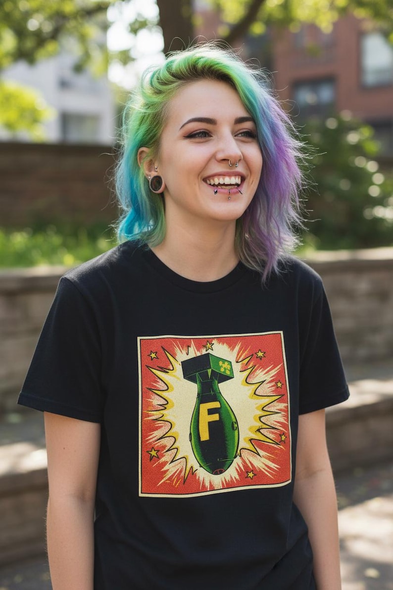 Pode incluir: Camiseta preta com um gr&aacute;fico de uma bomba com a letra "F" dentro de uma explos&atilde;o amarela, emoldurada por uma borda vermelha com estrelas. A pessoa que usa a camiseta tem cabelo multicolorido.