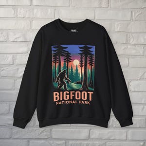 May include: Black sweatshirt featuring a Bigfoot National Park design. The graphic depicts a Bigfoot silhouette walking through a forest at sunset, with the words "BIGFOOT NATIONAL PARK" in orange. The design is centered on the front of the sweatshirt.