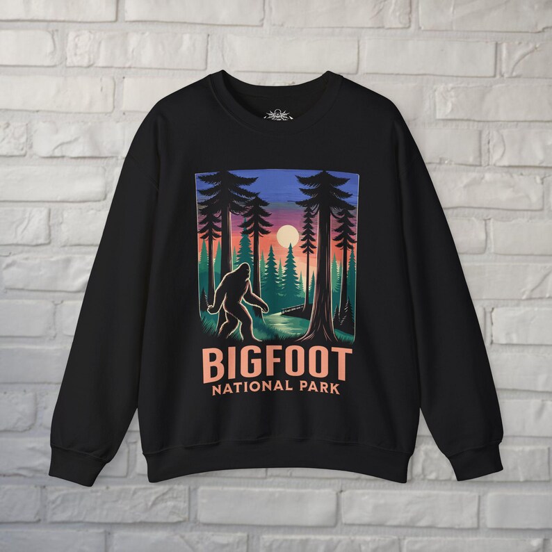 May include: Black sweatshirt featuring a Bigfoot National Park design. The graphic depicts a Bigfoot silhouette walking through a forest at sunset, with the words "BIGFOOT NATIONAL PARK" in orange. The design is centered on the front of the sweatshirt.