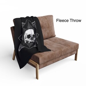 May include: Black fleece throw blanket featuring a white skull design. The skull is centered within a geometric starburst pattern. The blanket is draped over a brown upholstered chair. The text "Fleece Throw" is visible in the image.