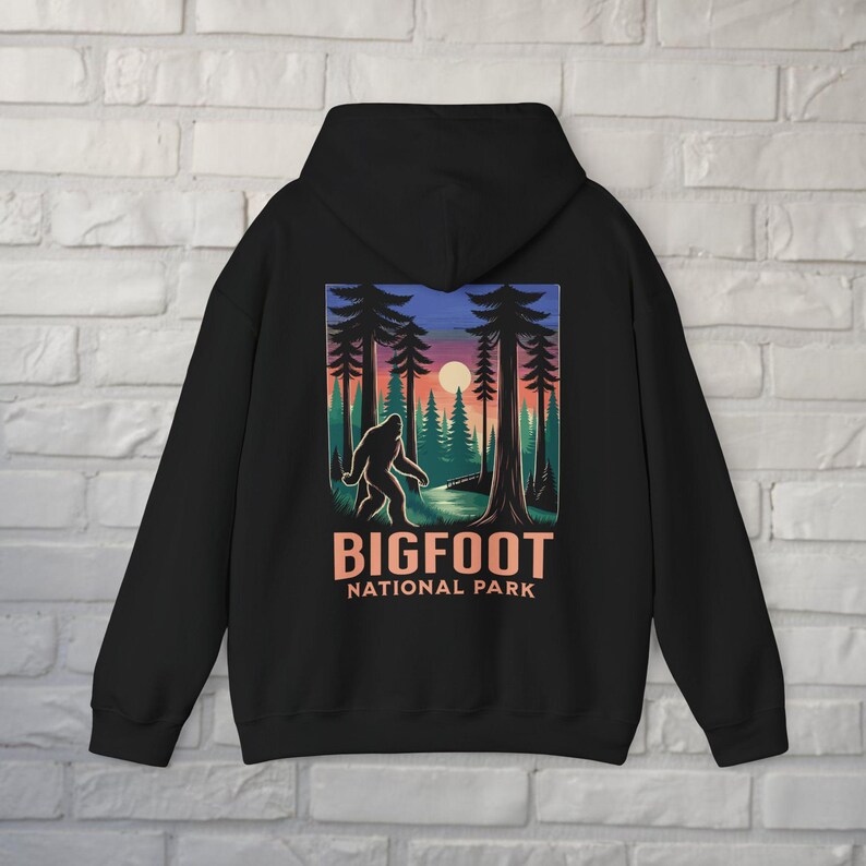 May include: Black hooded sweatshirt featuring a Bigfoot graphic in a forest scene with a sunset. The design includes the text "BIGFOOT NATIONAL PARK" in orange and white. The sweatshirt has a drawstring hood.