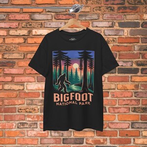 May include: Black t-shirt featuring a Bigfoot graphic in a forest scene with a sunset. The design includes the text "BIGFOOT NATIONAL PARK" in a stylized font. The shirt is hanging on a wooden hanger.