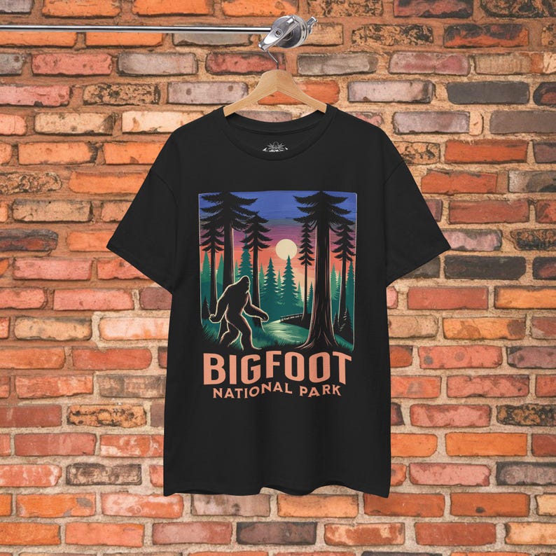May include: Black t-shirt featuring a Bigfoot graphic in a forest scene with a sunset. The design includes the text "BIGFOOT NATIONAL PARK" in a stylized font. The shirt is hanging on a wooden hanger.