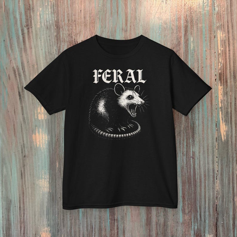 Puede incluir: Una camiseta negra de manga corta con una ilustraci&oacute;n gr&aacute;fica blanca de una zarig&uuml;eya siseando centrada en el pecho. Sobre la zarig&uuml;eya, la palabra FERAL est&aacute; impresa en una fuente g&oacute;tica blanca y negrita. El dise&ntilde;o utiliza tinta de alto contraste.