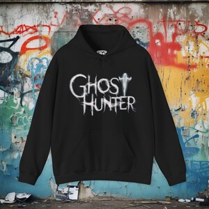 May include: A black hooded sweatshirt featuring a white graphic on the chest that reads GHOST HUNTER in a stylized, wispy font. The design includes a small, ethereal figure standing above the text. The hoodie has long sleeves and a front pocket.