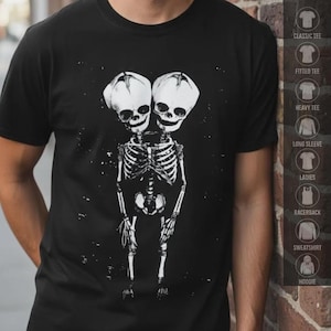 May include: A black short-sleeve t-shirt featuring a white graphic print of a two-headed skeleton figure. The skeleton has two distinct skulls atop a single ribcage and pelvic structure, with arms hanging down at the sides against a dark background.