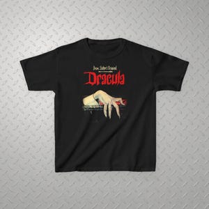 May include: A black short-sleeve t-shirt featuring a graphic print of a pale hand with long fingernails resting on a dark ledge. Above the hand, the text reads Bram Stoker's Original Dracula in a stylized red and white font on the chest.