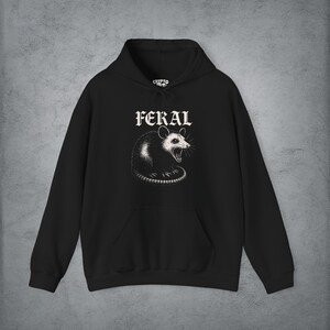 Puede incluir: Una sudadera con capucha negra que presenta un gr&aacute;fico blanco de una zarig&uuml;eya siseando centrado en el pecho. Sobre la zarig&uuml;eya, la palabra FERAL est&aacute; impresa en una fuente g&oacute;tica blanca. La prenda incluye un bolsillo canguro y mangas largas.