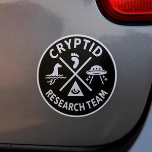 May include: A round black and white Cryptid Research Team sticker. The sticker features a Nessie-like creature, a footprint, a UFO, and an all-seeing eye. The words "CRYPTID" and "RESEARCH TEAM" are in a circular design.