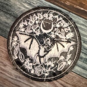 May include: A black and white embroidered patch featuring a bat with outstretched wings, a crescent moon, and a floral design. The patch is circular and has a white border.