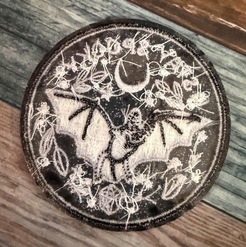 May include: A black and white embroidered patch featuring a bat with outstretched wings, a crescent moon, and a floral design. The patch is circular and has a white border.