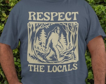 Może przedstawiać: Niebieska koszulka z grafiką przedstawiającą Bigfoota idącego przez las. Pod grafiką znajduje się napis "Respect the Locals".