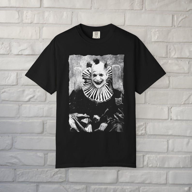 May include: A black short-sleeve t-shirt featuring a centered, high-contrast black and white graphic print of a clown with a wide, dark smile and a ruffled collar. The clown is depicted in a seated position, rendered in a gritty, textured style.
