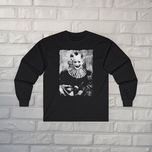 May include: A black long-sleeve shirt featuring a centered, high-contrast black and white graphic print of a clown with a wide, toothy grin and a ruffled collar. The design has a distressed, textured appearance, set against the solid black fabric.