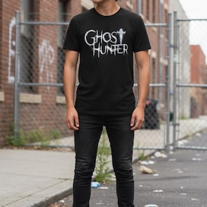 May include: A black short-sleeved t-shirt featuring the white text GHOST HUNTER in a stylized, distressed font across the chest, paired with black slim-fit jeans and black boots, creating a monochromatic outfit with a graphic design element.