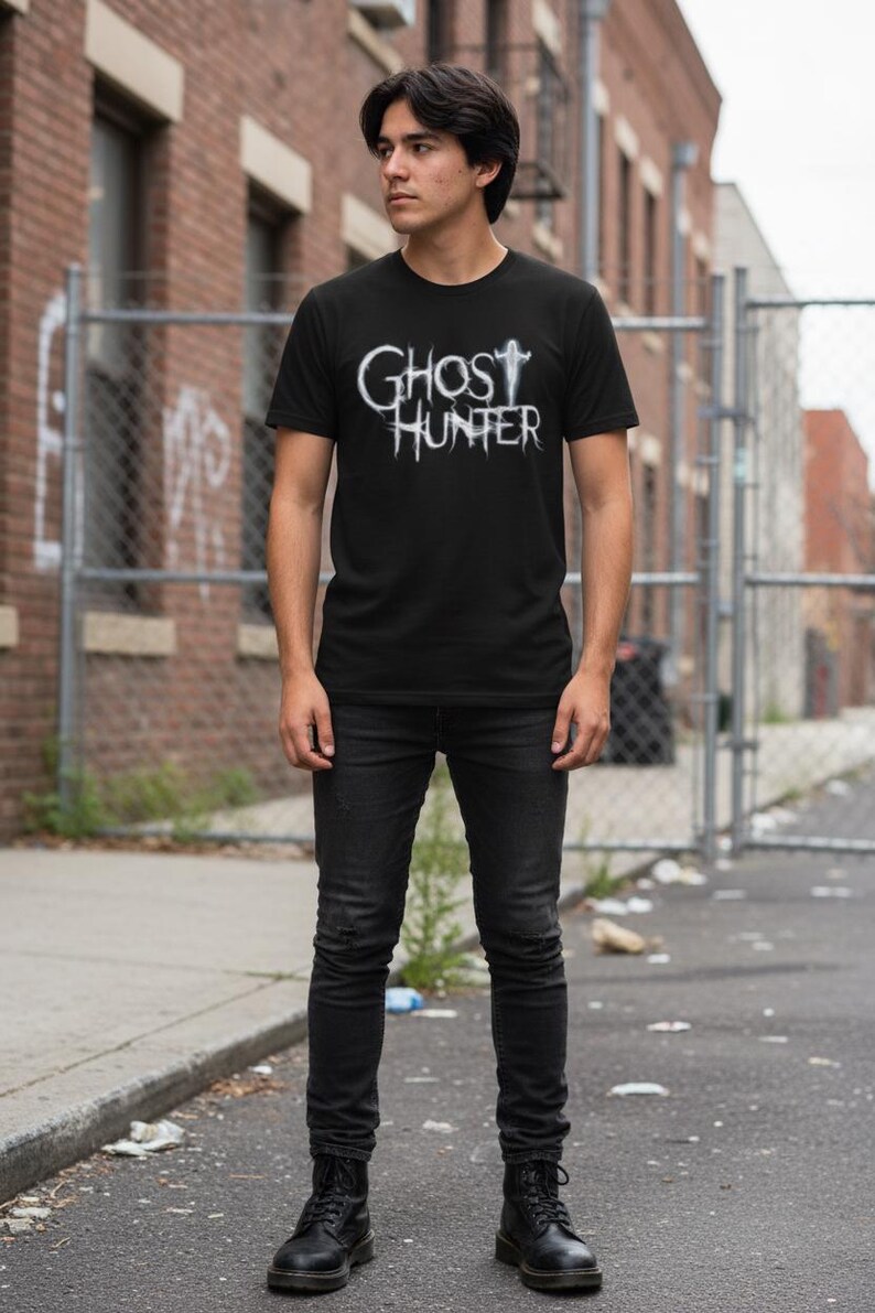 May include: A black short-sleeved t-shirt featuring the white text GHOST HUNTER in a stylized, distressed font across the chest, paired with black slim-fit jeans and black boots, creating a monochromatic outfit with a graphic design element.