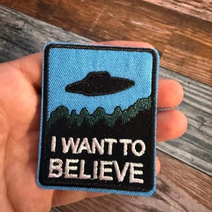 May include: A blue and black embroidered patch with a UFO and the text "I WANT TO BELIEVE".