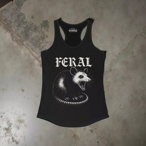 Puede incluir: Una camiseta de tirantes negra con espalda nadadora que presenta un estampado gr&aacute;fico blanco de una zarig&uuml;eya siseando. La palabra FERAL est&aacute; impresa en una fuente g&oacute;tica llamativa sobre la ilustraci&oacute;n. Incluye un peque&ntilde;o logo en el cuello.