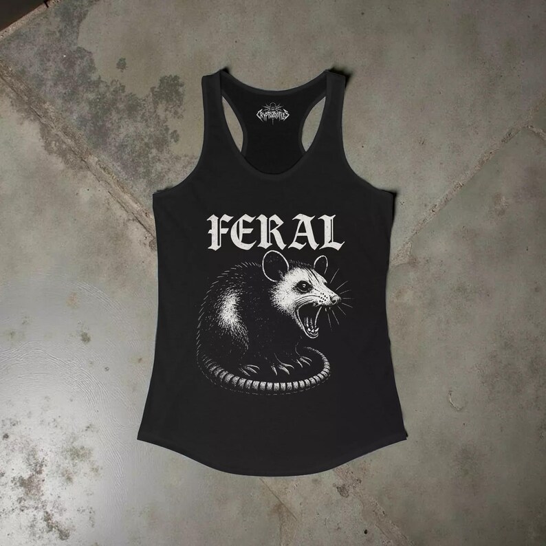 Puede incluir: Una camiseta de tirantes negra con espalda nadadora que presenta un estampado gr&aacute;fico blanco de una zarig&uuml;eya siseando. La palabra FERAL est&aacute; impresa en una fuente g&oacute;tica llamativa sobre la ilustraci&oacute;n. Incluye un peque&ntilde;o logo en el cuello.