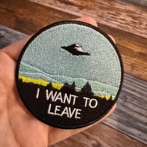 May include: Round embroidered patch with a black border. The patch features a light blue sky with a flying saucer, a black landscape, and the white text "I WANT TO LEAVE".
