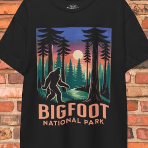 May include: Black t-shirt featuring a Bigfoot graphic in a forest scene with a sunset. The design includes the text "BIGFOOT NATIONAL PARK" in a stylized font. The illustration depicts a silhouette of Bigfoot walking through a forest.