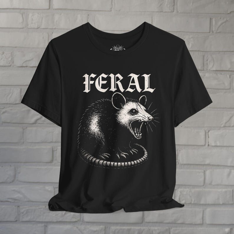Puede incluir: Una camiseta negra de manga corta con una ilustraci&oacute;n gr&aacute;fica blanca de una zarig&uuml;eya siseando. Sobre la zarig&uuml;eya, la palabra FERAL est&aacute; impresa en una fuente blanca, negrita y estilizada, centrada en la parte frontal de la prenda.