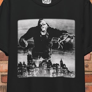 Peut inclure: T-shirt noir et blanc avec un grand gorille debout devant un horizon de ville. Le gorille tient une femme dans sa main.