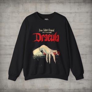 May include: A black long-sleeve sweatshirt featuring a graphic print on the chest. The text reads Bram Stoker's Original Dracula in red and white lettering. Below the text is an illustration of a pale hand with long fingernails resting on a ledge.
