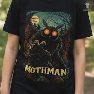 Puede incluir: Una camiseta negra con un estampado gráfico del Mothman con ojos naranjas brillantes, alas grandes y una mano con garras extendida hacia adelante. El fondo muestra un bosque y una luna llena, con el texto MOTHMAN impreso en amarillo en la parte inferior.