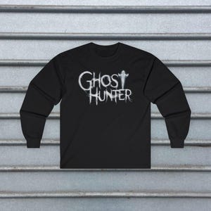 May include: A black long-sleeve shirt featuring the white text GHOST HUNTER printed on the chest. The lettering has a wispy, ethereal appearance, with the letter T in GHOST stylized to resemble a faint, glowing silhouette of a human figure.