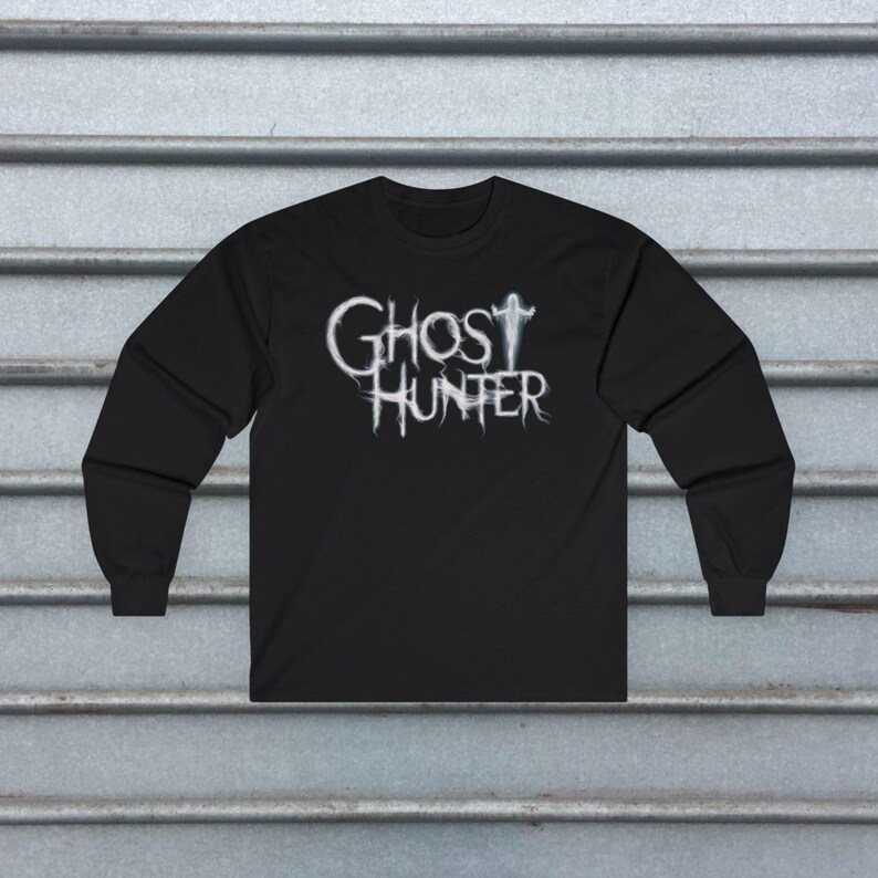 May include: A black long-sleeve shirt featuring the white text GHOST HUNTER printed on the chest. The lettering has a wispy, ethereal appearance, with the letter T in GHOST stylized to resemble a faint, glowing silhouette of a human figure.