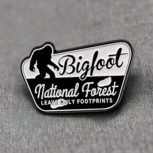 May include: Black and white enamel pin with a silhouette of Bigfoot and the text "Bigfoot National Forest Leave Only Footprints".