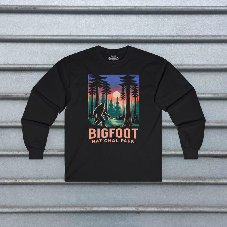 May include: Black long-sleeve shirt featuring a Bigfoot graphic in a forest scene with a sunset. The design includes the text "BIGFOOT NATIONAL PARK" in a bold, orange font. The shirt is made of a soft material.