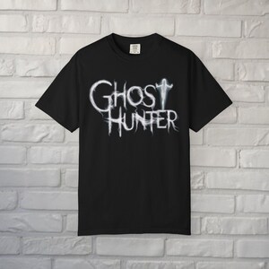 May include: A black short-sleeved t-shirt featuring the white text Ghost Hunter in a wispy, smoke-like font. The letter T in Ghost is stylized as a small, glowing white figure with arms outstretched. The shirt has a crew neckline and a standard fit.