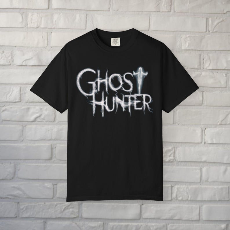 May include: A black short-sleeved t-shirt featuring the white text Ghost Hunter in a wispy, smoke-like font. The letter T in Ghost is stylized as a small, glowing white figure with arms outstretched. The shirt has a crew neckline and a standard fit.