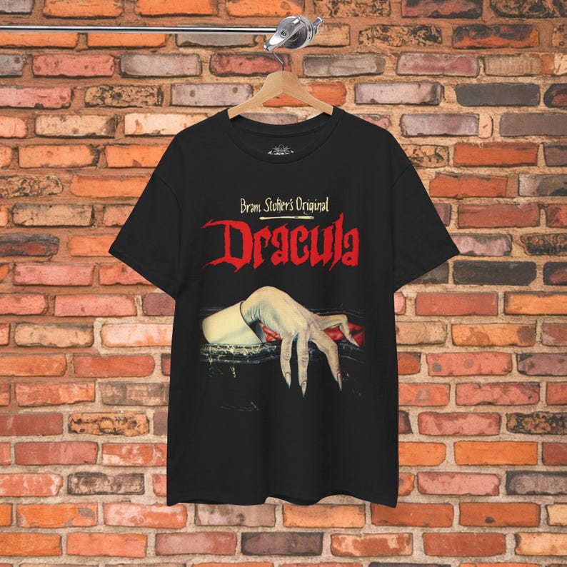 May include: A black short-sleeve t-shirt featuring the text Bram Stoker's Original Dracula in red and white lettering. Below the text is a graphic illustration of a pale, claw-like hand emerging from a dark, rectangular opening with red fabric.