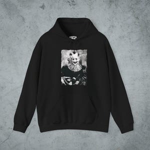 May include: A black hooded sweatshirt featuring a central graphic print of a clown with a wide smile and a ruffled collar. The design is rendered in a high-contrast black and white style. The hoodie includes a front kangaroo pocket and a drawstring.