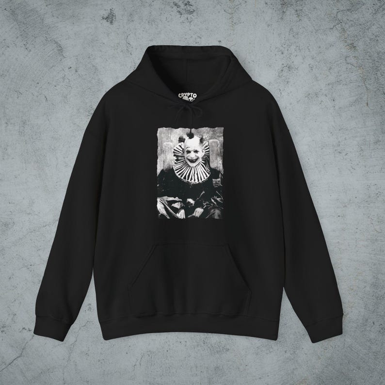 May include: A black hooded sweatshirt featuring a central graphic print of a clown with a wide smile and a ruffled collar. The design is rendered in a high-contrast black and white style. The hoodie includes a front kangaroo pocket and a drawstring.