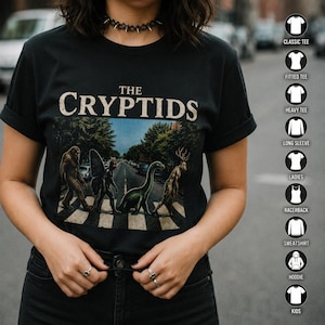 May include: A black t-shirt featuring the text THE CRYPTIDS above a graphic of four creatures walking across a crosswalk. The creatures include a Bigfoot, a Mothman, a Loch Ness Monster, and a Jackalope. A column of shirt style icons is on the right.
