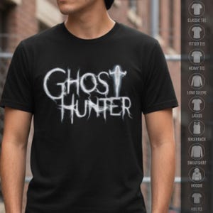 May include: A black short-sleeved t-shirt featuring the white text GHOST HUNTER in a stylized, wispy font that resembles smoke or mist. The letter T in GHOST is replaced by a small, glowing white silhouette of a figure with arms outstretched.
